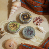 Three small metal tins with decorative labels on a sandy surface with shells and a towel.