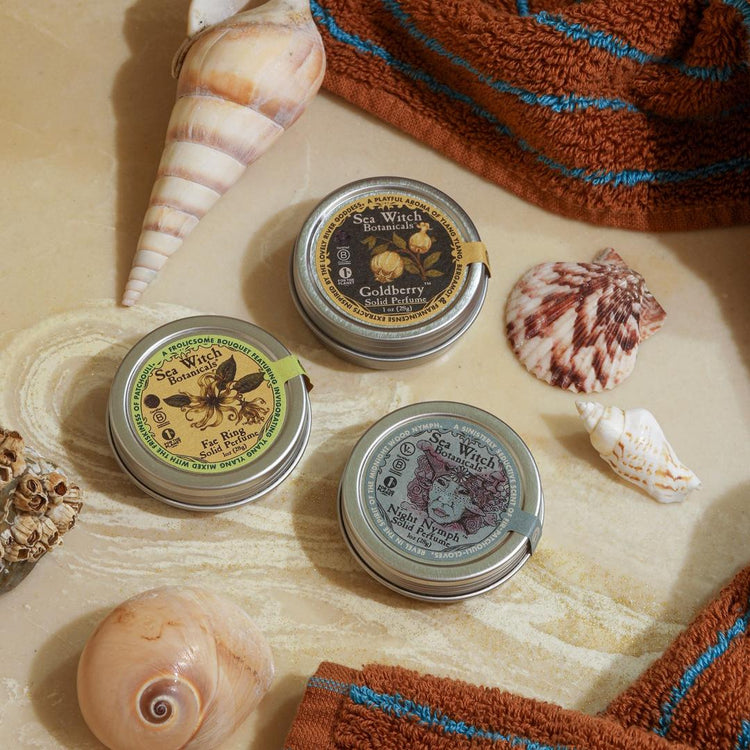 Three small metal tins with decorative labels on a sandy surface with shells and a towel.